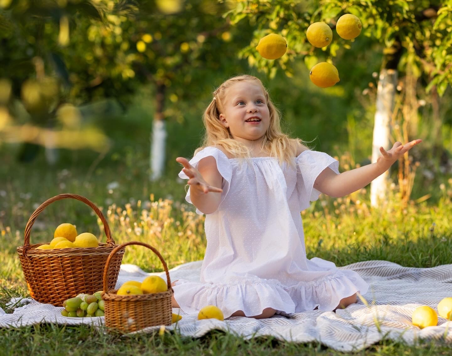Uśmiechnięta blond dziewczynka w białej sukience żongluje cytrynami na pikniku w sadzie. Kosze z owocami, miękkie światło, radosna atmosfera.