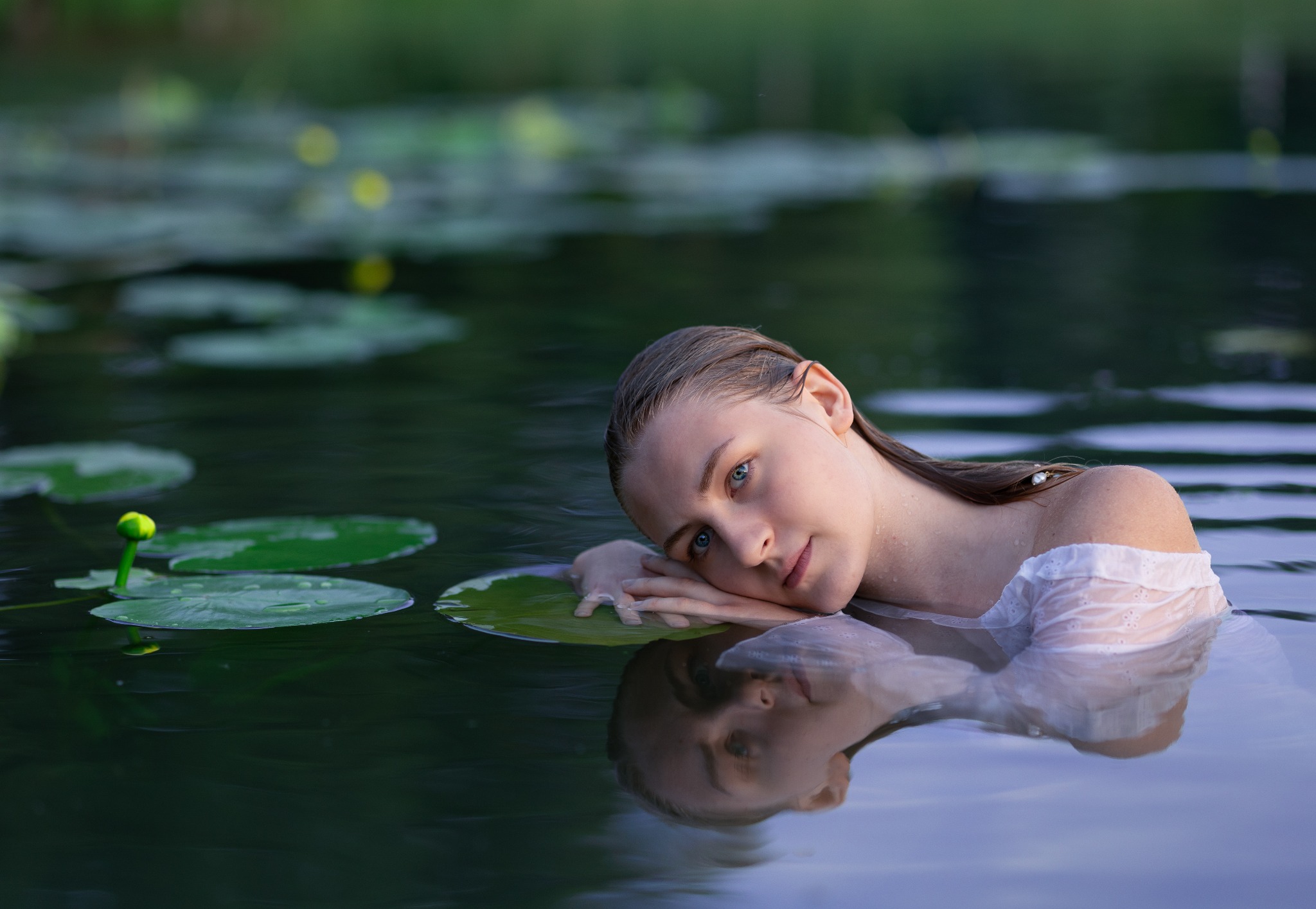 Portret kobiety w wodzie, opierającej się o liść lilii wodnej. Delikatne światło, spokojna atmosfera, naturalne otoczenie. Widoczne odbicie w wodzie.