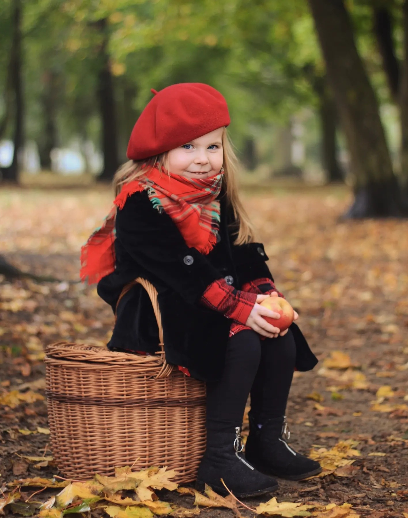Uśmiechnięta dziewczynka w czerwonym berecie i szaliku siedzi na wiklinowym koszu w jesiennym parku, trzymając jabłko. Ubrana w czarny płaszczyk i czarne buty.