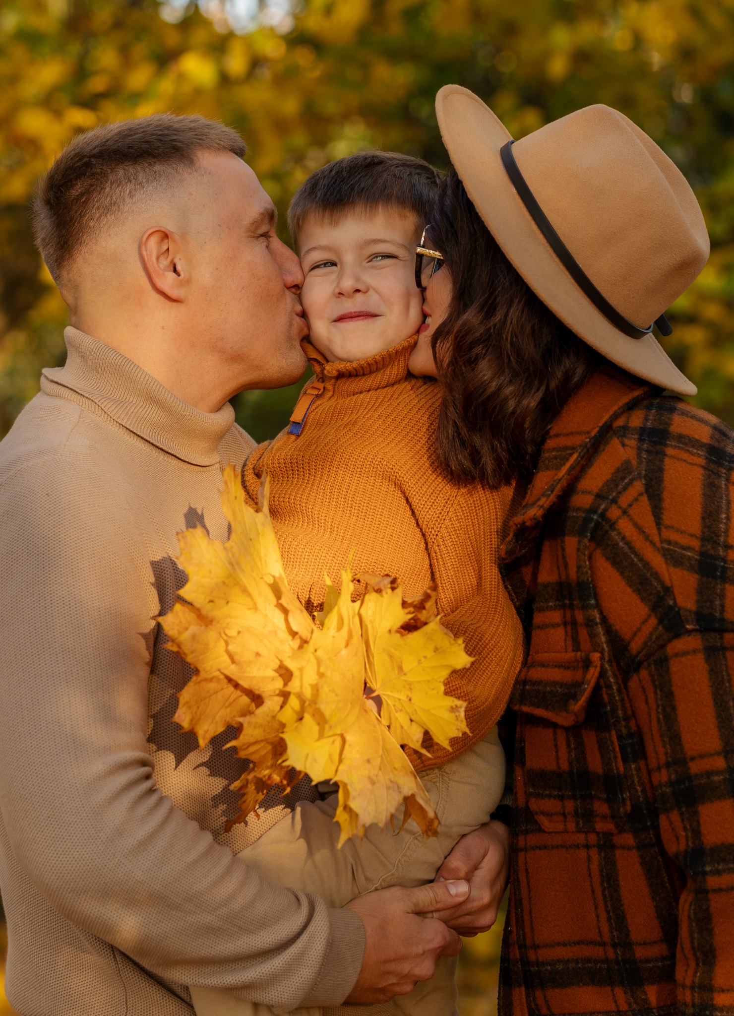 Jesienna sesja rodzinna: rodzice całują uśmiechniętego syna trzymającego bukiet żółtych liści na tle drzew w parku, ciepłe kolory, miękkie światło.