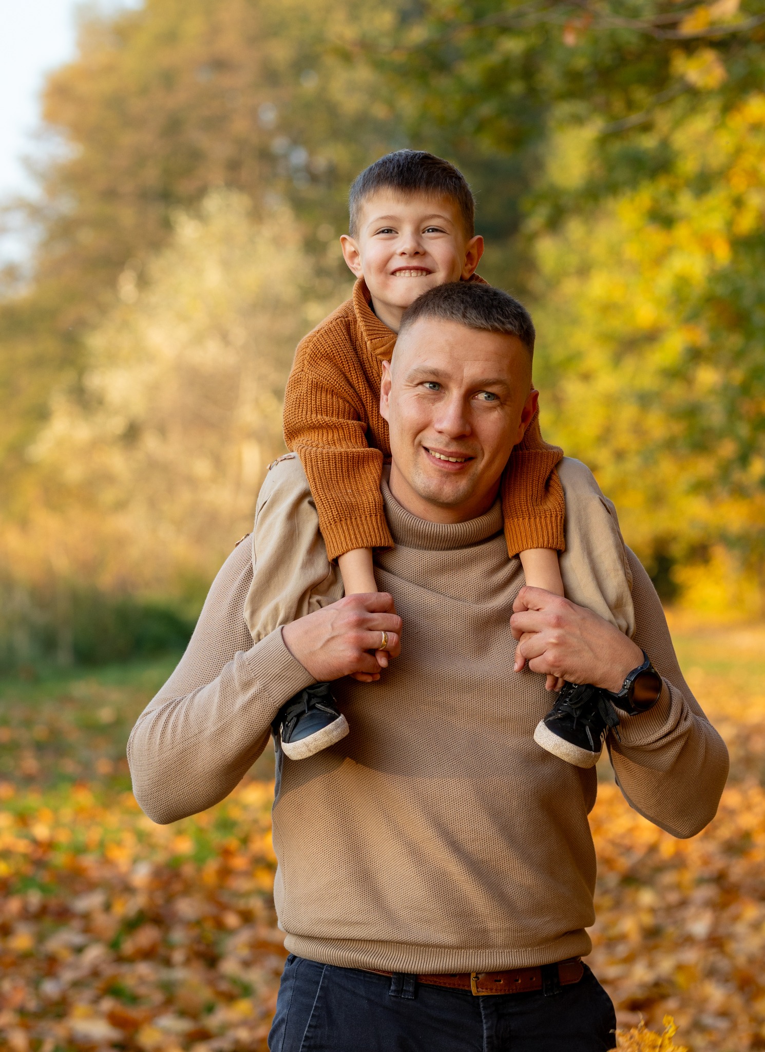Uśmiechnięty ojciec z synem na barana w jesiennym parku. Ciepłe, naturalne światło podkreśla więź. Widoczne opadłe liście i drzewa w tle.