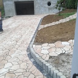 Podhale Invest Bud - Kremowa ścieżka z nieregularnych płyt prowadzi do garażu z okrągłym oknem. Obrzeże z ciemnych kostek i betonowych palisad. Część ścieżki jeszcze nieukończona.