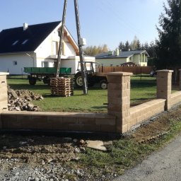 Podhale Invest Bud - Wznoszony, niski murek z beżowych bloczków betonowych, tworzący ogrodzenie posesji. W tle dom, traktor i słupy energetyczne. Widoczne ślady prac ziemnych.