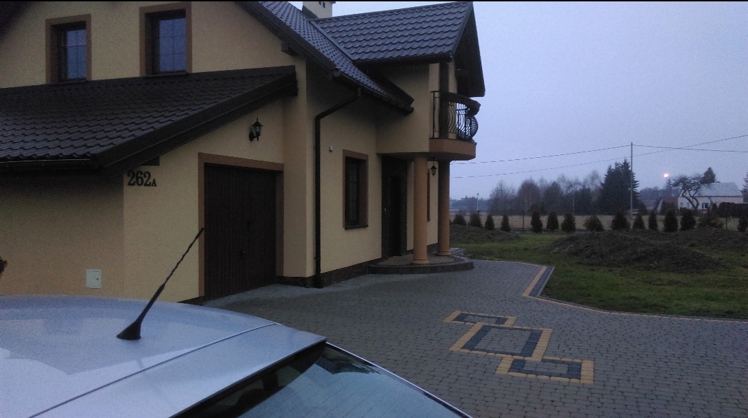 Dwukondygnacyjny dom z beżową elewacją, ciemnym dachem i balkonem. Na pierwszym planie fragment samochodu, a obok domu ułożona kostka brukowa z dekoracyjnym wzorem.