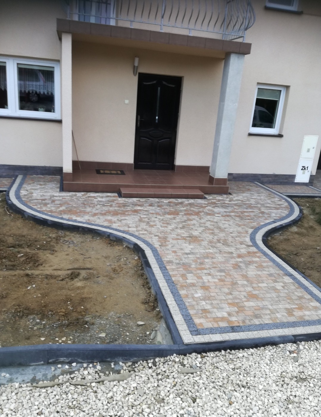 Ułożona kostka brukowa tworząca ścieżkę do drzwi wejściowych domu. Widoczne obrzeża i fragmenty niezabudowanego terenu. Jasna elewacja budynku w tle.