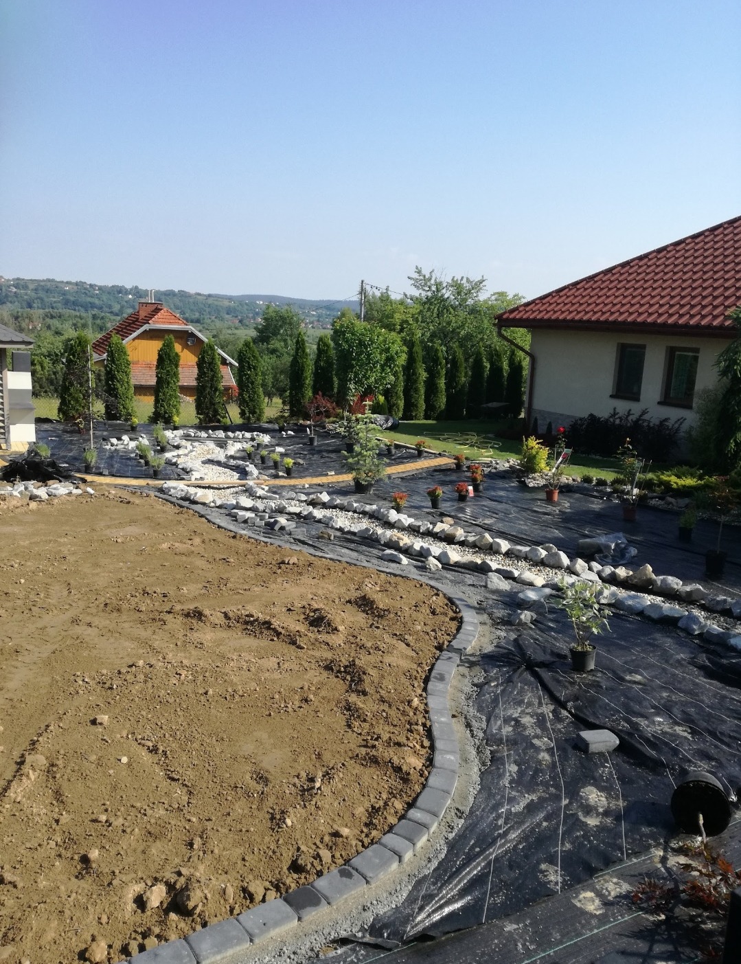 Aranżacja ogrodu w trakcie realizacji: ułożona agrowłóknina, obrzeża z kostki, kamienie ozdobne i sadzonki roślin na tle domów i zieleni w słoneczny dzień.