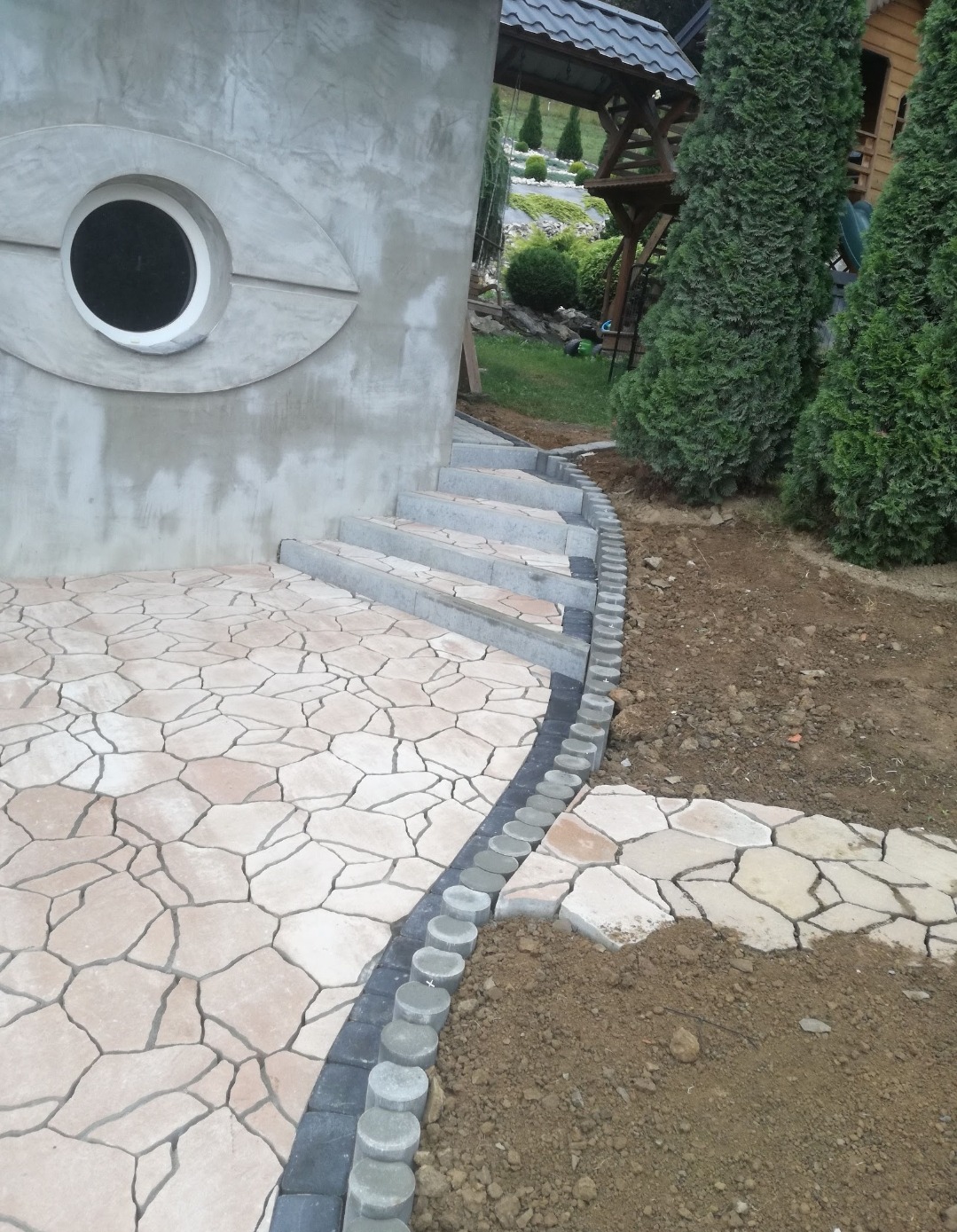 Ukończona ścieżka z kostki brukowej o nieregularnym kształcie, obrzeża z szarej kostki. Schody z jasnego kamienia prowadzą do budynku z oknem w kształcie oka.