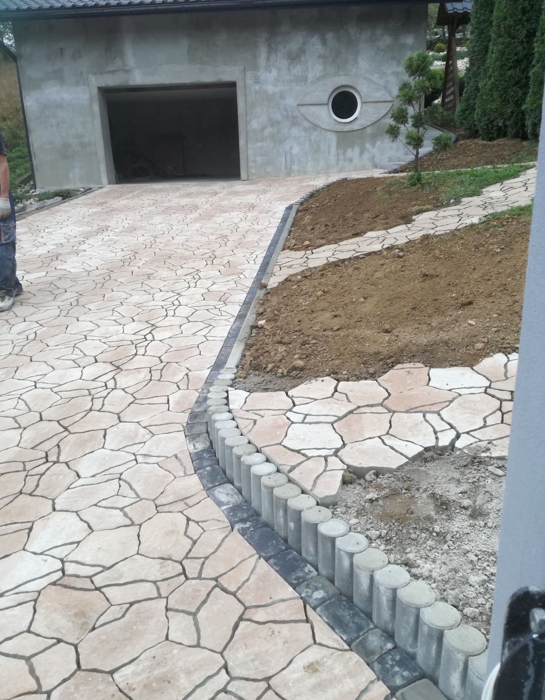 Kremowa ścieżka z nieregularnych płyt prowadzi do garażu z okrągłym oknem. Obrzeże z ciemnych kostek i betonowych palisad. Część ścieżki jeszcze nieukończona.