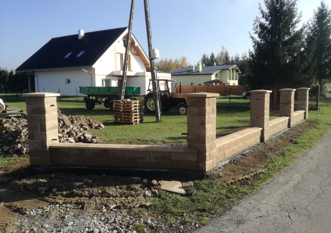 Wznoszony, niski murek z beżowych bloczków betonowych, tworzący ogrodzenie posesji. W tle dom, traktor i słupy energetyczne. Widoczne ślady prac ziemnych.