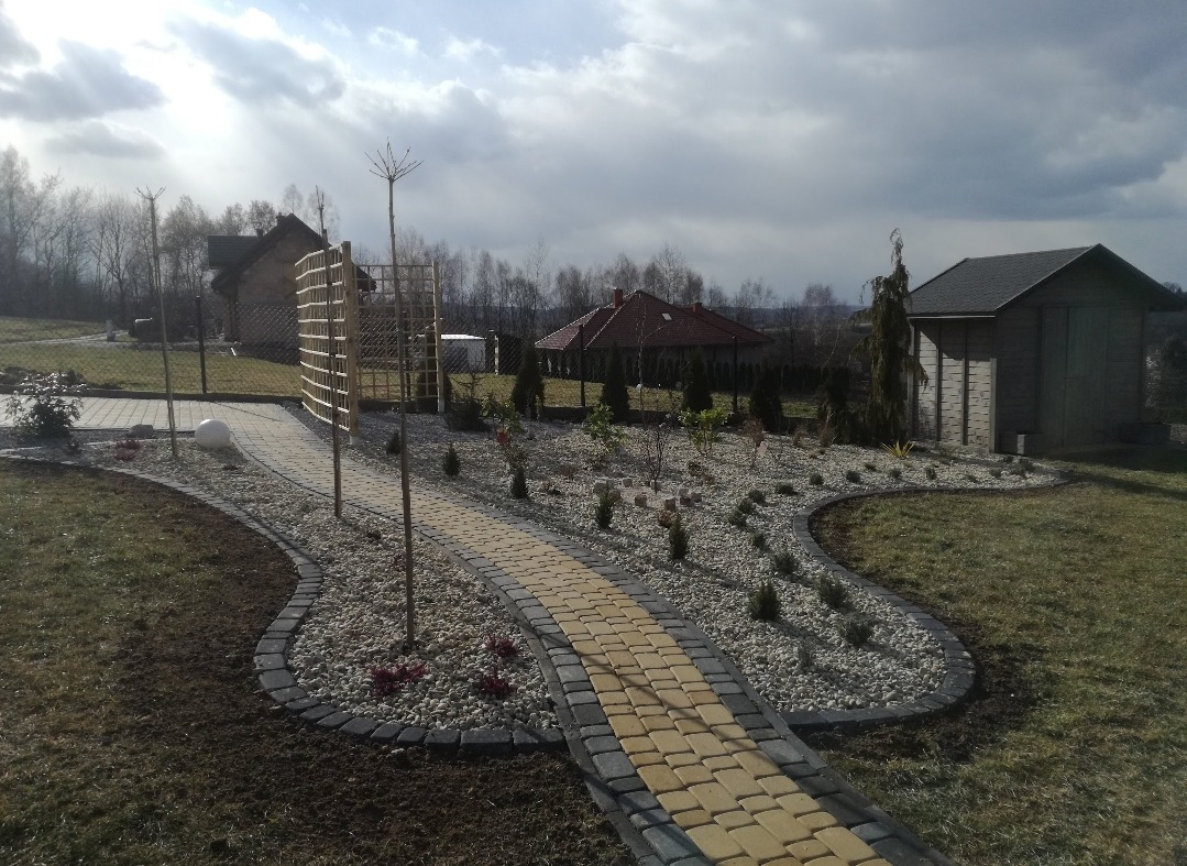 Żółta ścieżka z kostki brukowej, obrzeża z szarej kostki, rabaty z białym kamieniem i roślinami. W tle altana i budynek gospodarczy, pochmurne niebo.