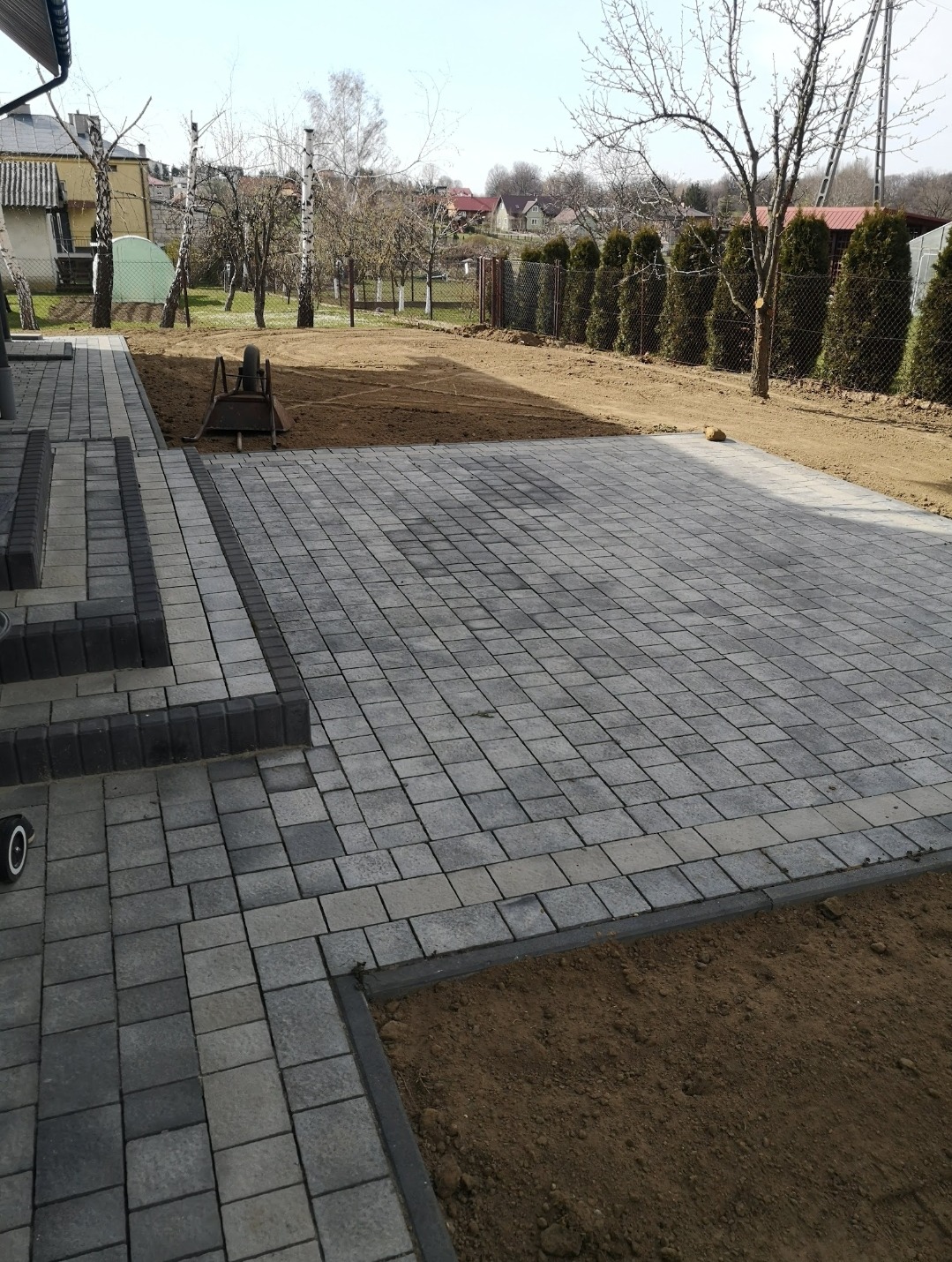 Ułożona kostka brukowa w odcieniach szarości tworząca plac przed domem. Widoczny fragment schodów i przygotowany teren pod dalsze prace. W tle drzewa i ogrodzenie.