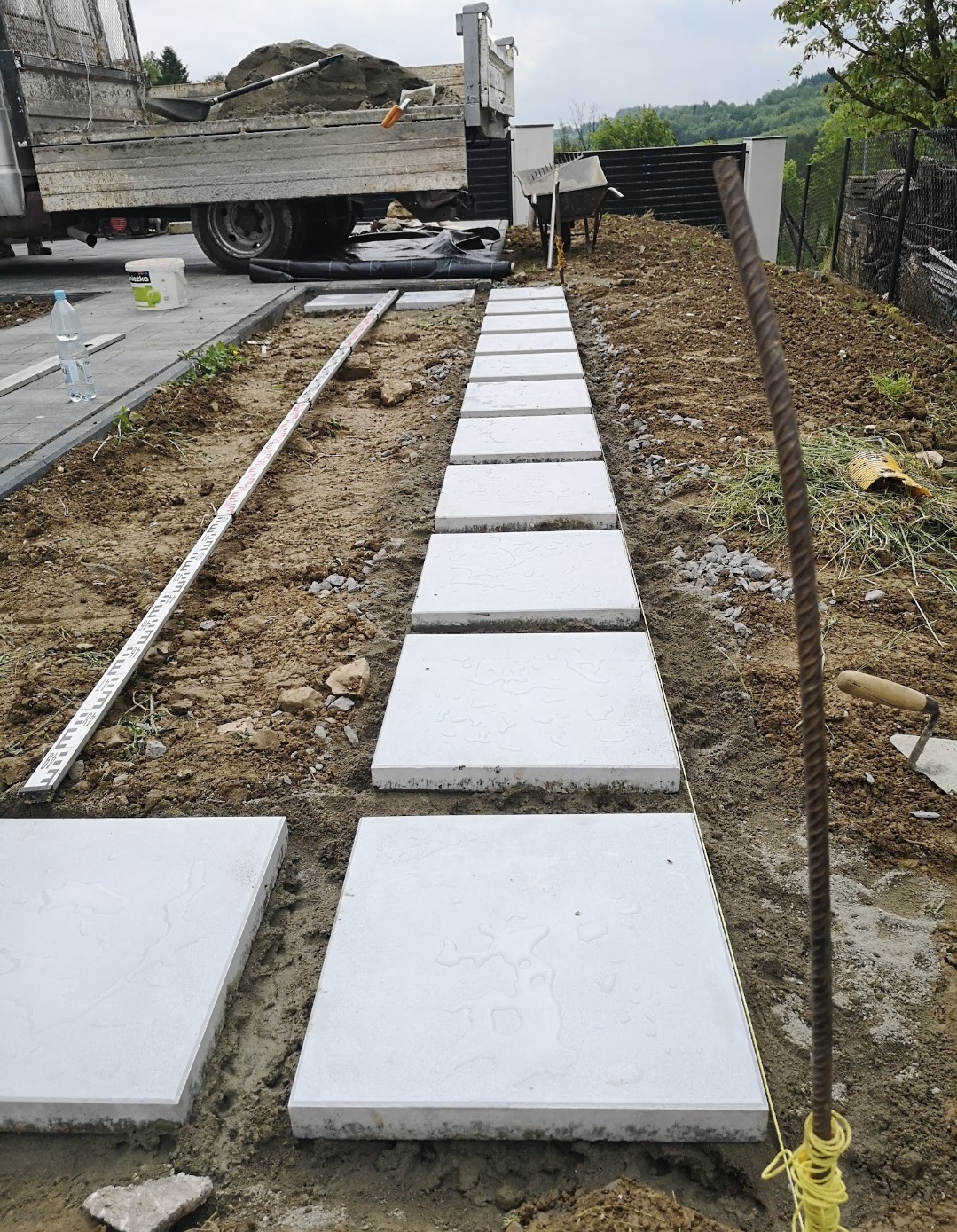 Ścieżka z jasnych, kwadratowych płyt ułożonych w ziemi, z widocznymi narzędziami i ciężarówką w tle. Etap prac brukarskich w ogrodzie.