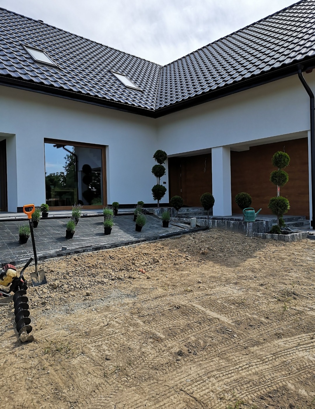 Przygotowanie terenu pod ogród: wykopana ziemia, narzędzia (wiertnica, łopata), kostka brukowa, sadzonki w doniczkach, nowoczesny dom z ciemną dachówką w tle.