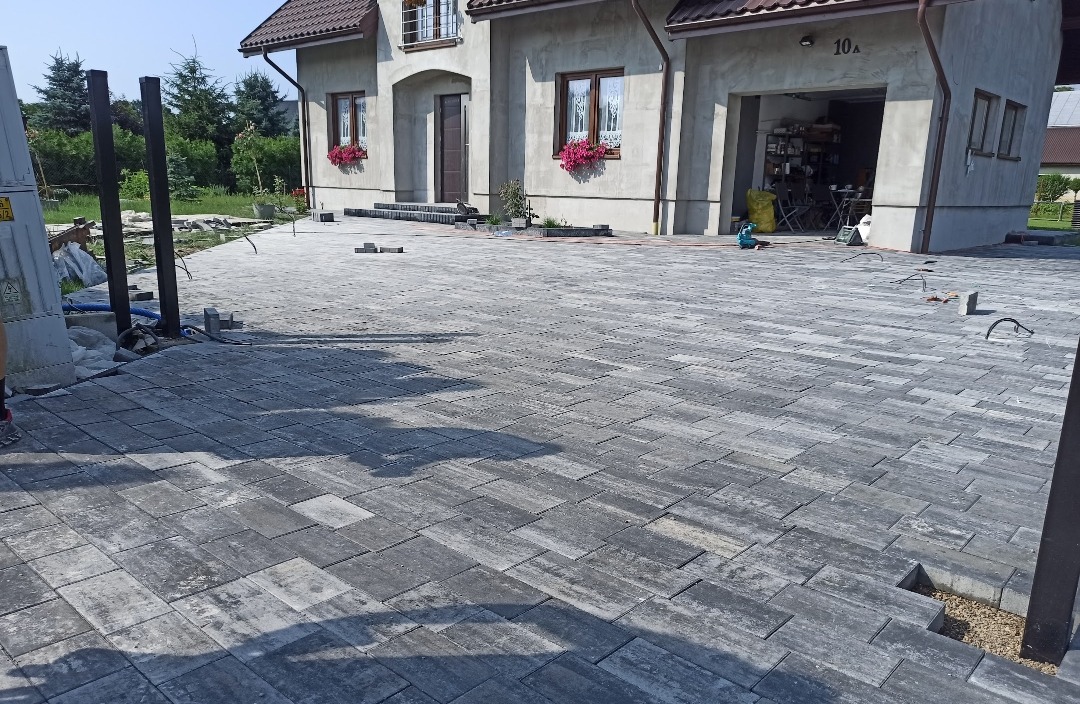 Ułożona kostka brukowa w szarym odcieniu przed domem z garażem i kwiatami w oknach. Częściowo widoczne słupki ogrodzeniowe i niezakończone fragmenty nawierzchni.