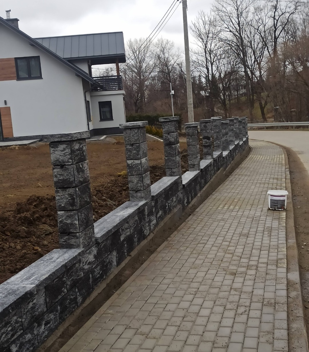 Nowoczesny, niski murek z szarej kostki łupanej, oddzielający posesję od chodnika z kostki. W tle dom z szarą stolarką i drzewa bez liści.