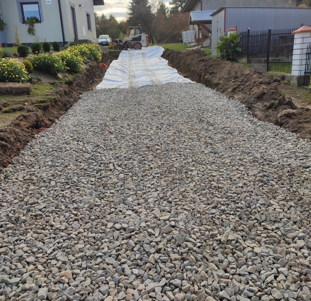 Utwardzony teren podjazdowy z warstwą tłucznia i geowłókniną, widoczny sprzęt budowlany w tle, dom jednorodzinny i fragment ogrodu. Sezon jesienny, pochmurne niebo.