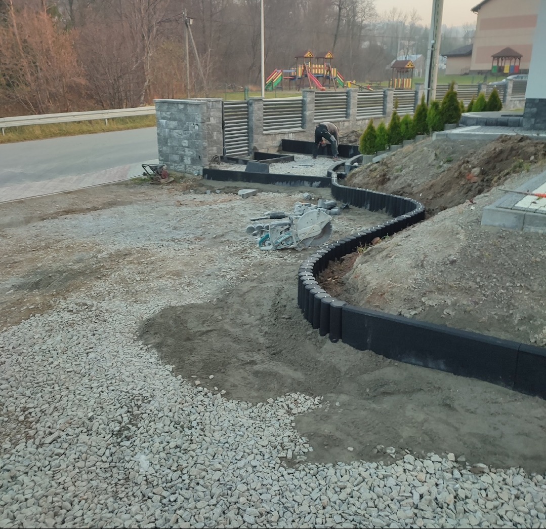 Krawężnik z palisady w ogrodzie, wyznaczający ścieżkę. W tle widoczny pracownik, narzędzia i plac zabaw. Palisada w kolorze czarnym, ułożona w łagodny łuk.