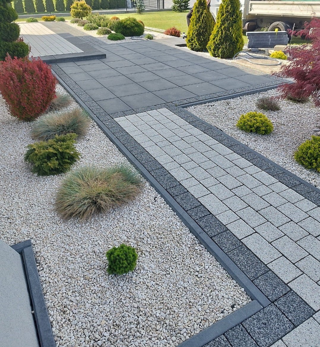 Nowoczesna aranżacja przestrzeni z kostki brukowej w odcieniach szarości, otoczona białym żwirem i zielenią. Geometryczny wzór ścieżki i podjazdu.