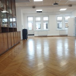 SPB CRYSTAL OLENA KRYCHUN - Wnętrze przestronnego pomieszczenia z parkietem ułożonym w jodełkę, po sprzątaniu. Widoczne okna, grzejniki, szafki wzdłuż ściany i projektor pod sufitem.