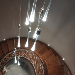 SPB CRYSTAL OLENA KRYCHUN - Spiralne schody z drewnianymi stopniami i metalową balustradą, oświetlone wiszącymi lampami z przezroczystego szkła, widziane z dołu, tworzące elegancki efekt wizualny.
