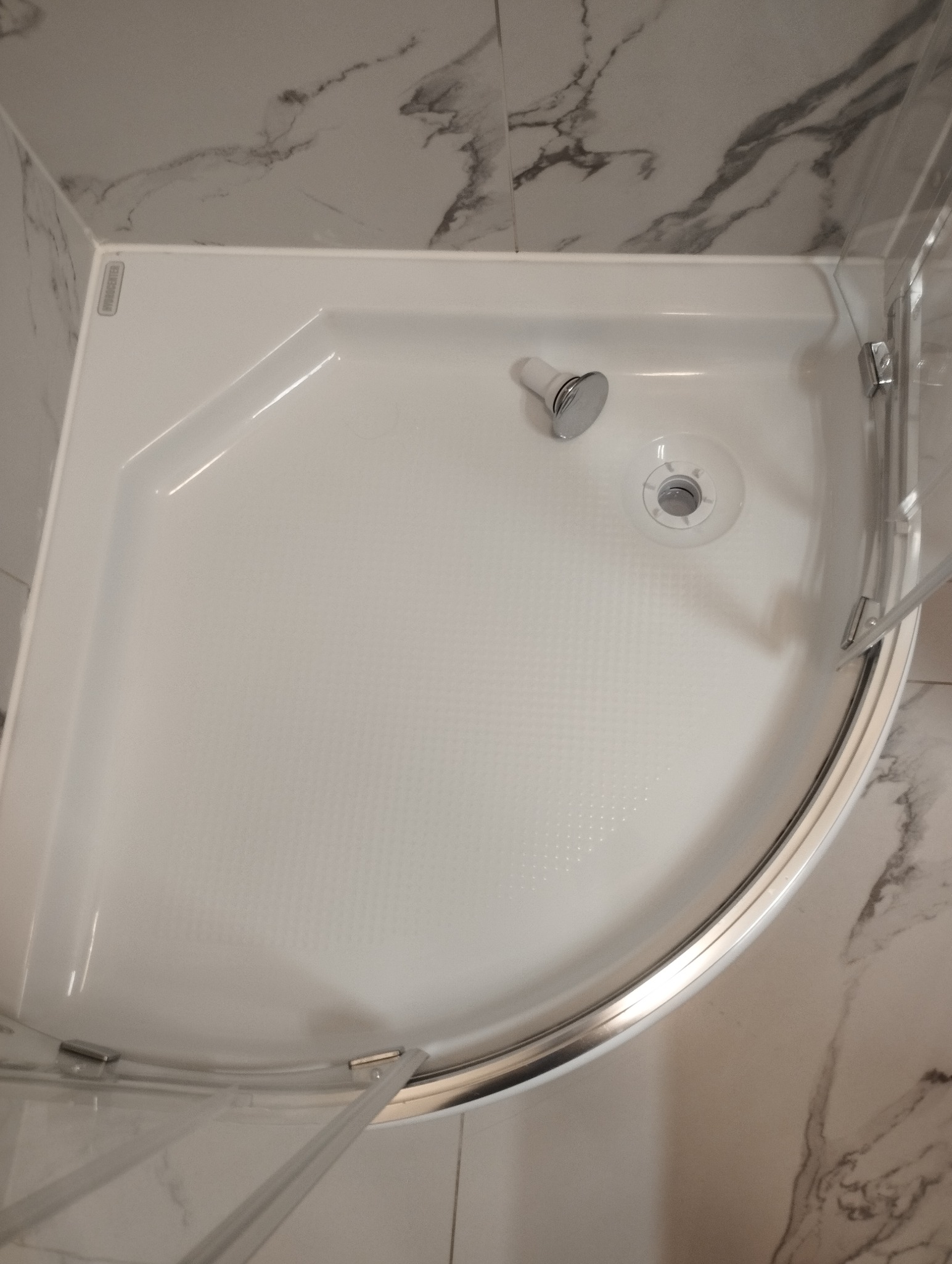 Biały brodzik kabiny prysznicowej z odpływem i chromowaną słuchawką, widok z góry. Marmurowe płytki na ścianach. Czystość i minimalizm.