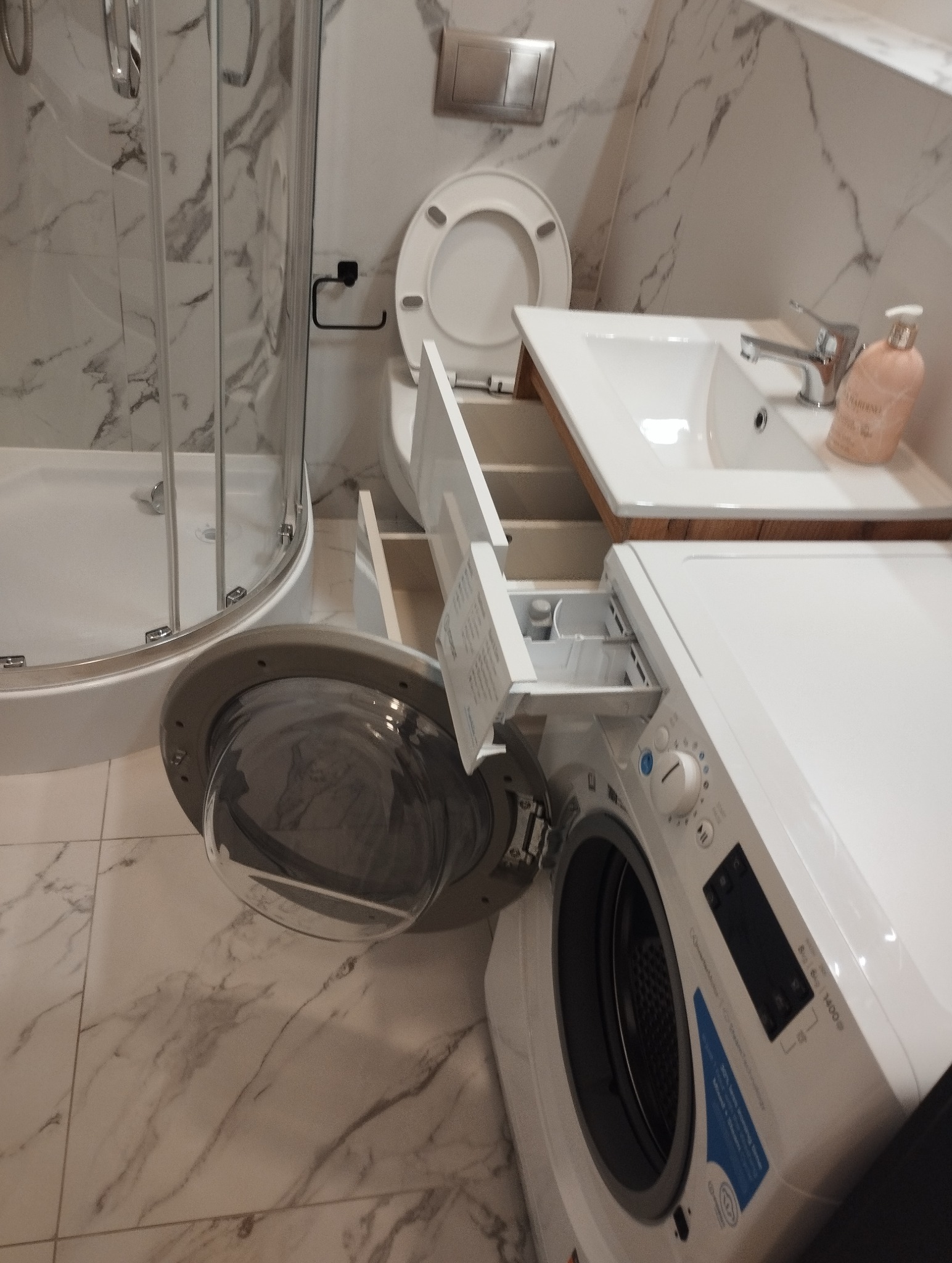 Biała pralka w łazience z marmurowymi płytkami, obok kabina prysznicowa i umywalka. Otwarty dozownik na proszek. Widok na czyste, błyszczące wnętrze łazienki.