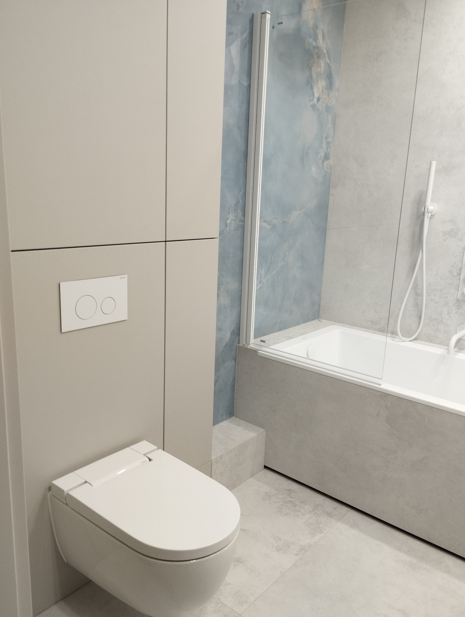 Nowoczesna łazienka w odcieniach szarości i błękitu z podwieszaną toaletą, zabudową meblową i wanną z parawanem. Minimalistyczny design, płytki imitujące kamień.