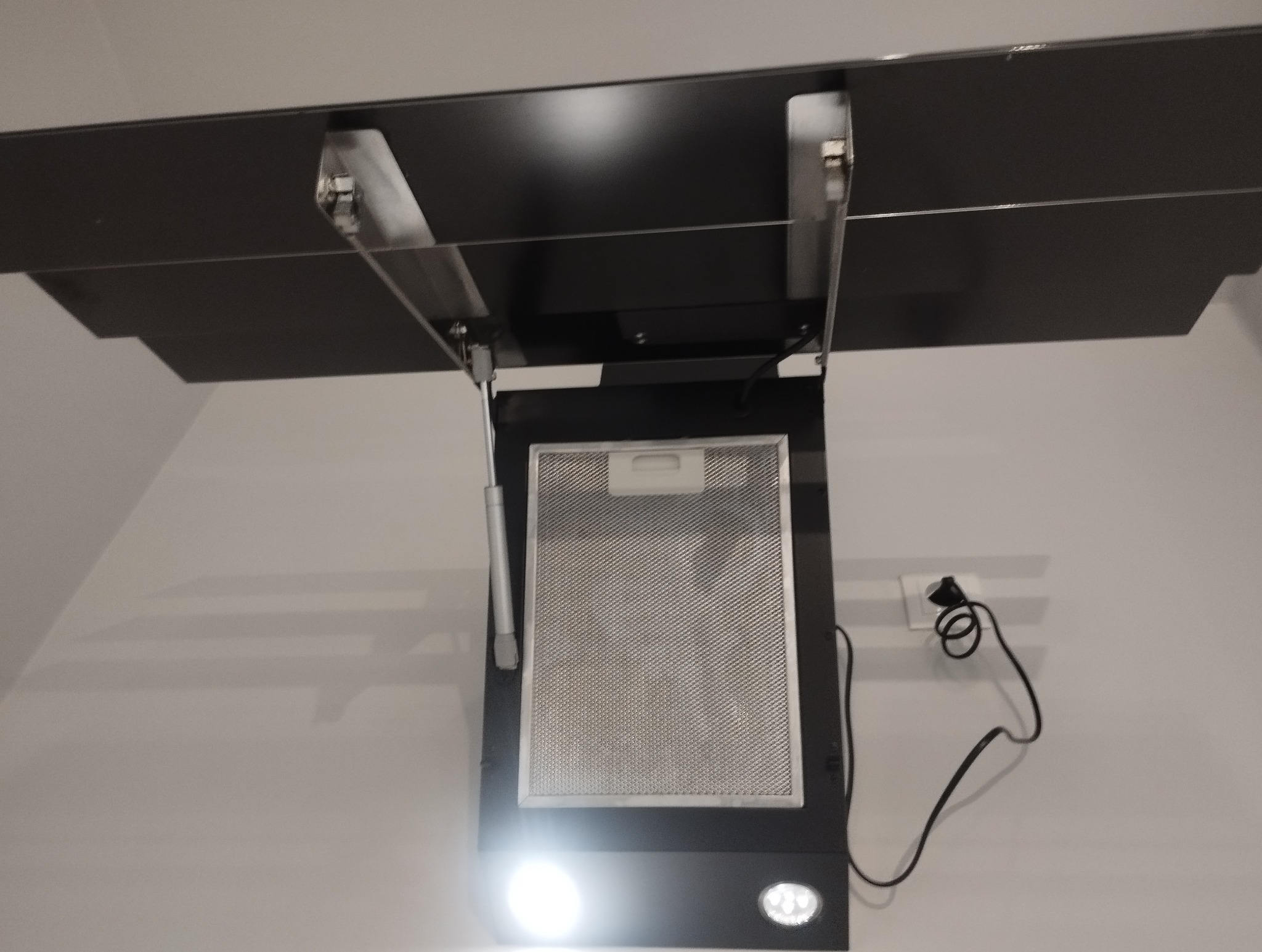Otwarty, czarny okap kuchenny z metalowym filtrem i włączonym oświetleniem, zamontowany na jasnej ścianie. Widoczny kabel zasilający i elementy mocujące.