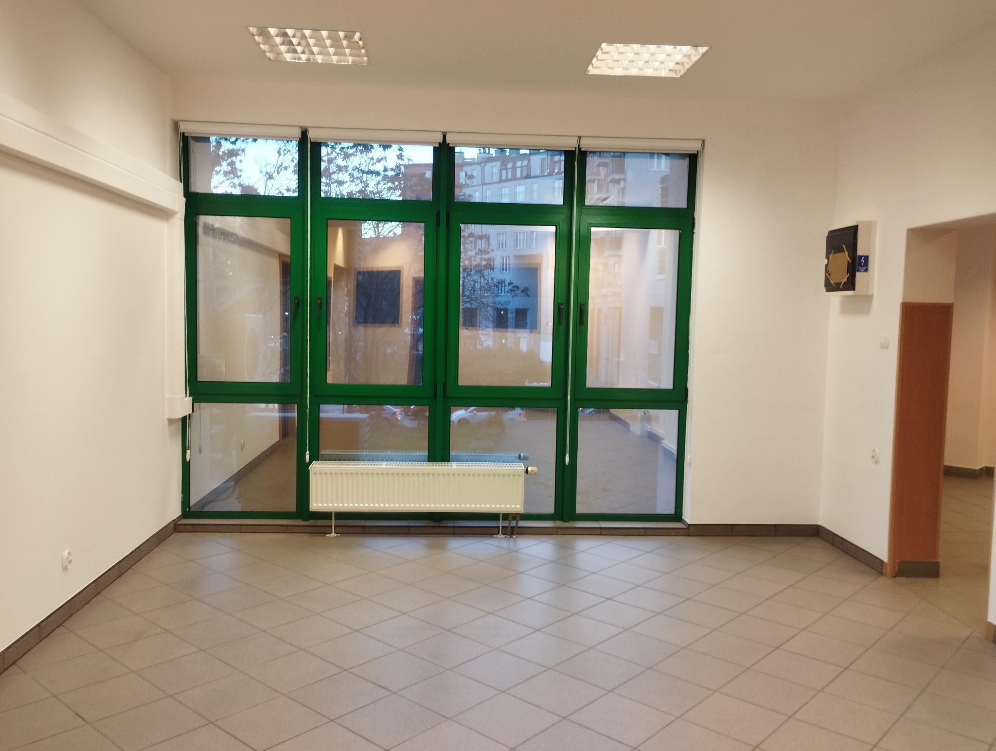 Puste, jasne pomieszczenie z dużymi oknami w zielonych ramach. Szara podłoga z płytek, białe ściany. Widoczny grzejnik pod oknem i fragment korytarza.