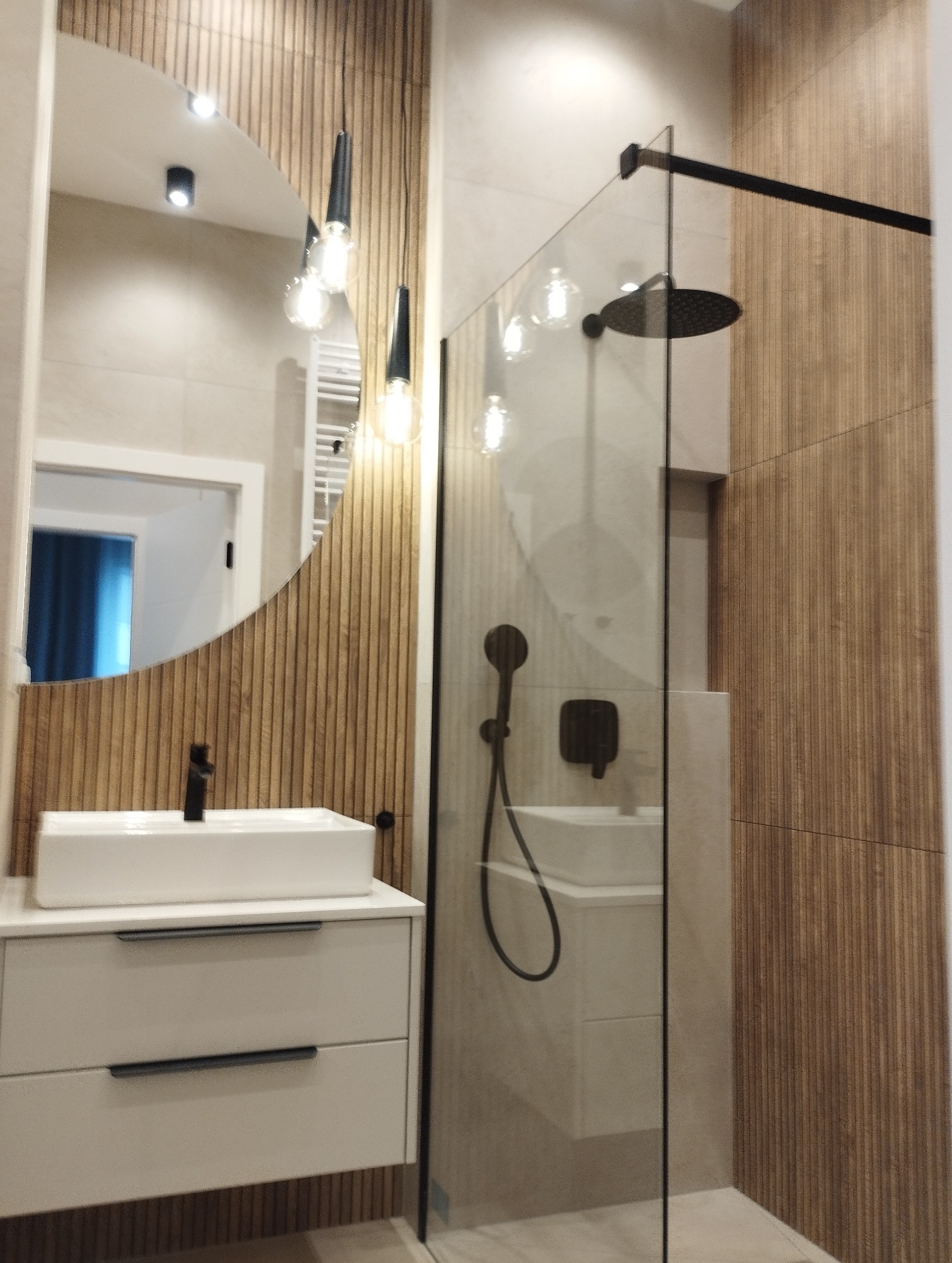 Nowoczesna łazienka po remoncie: kabina prysznicowa typu walk-in, umywalka nablatowa z szafką, okrągłe lustro z dekoracyjnym oświetleniem i drewniane panele ścienne.