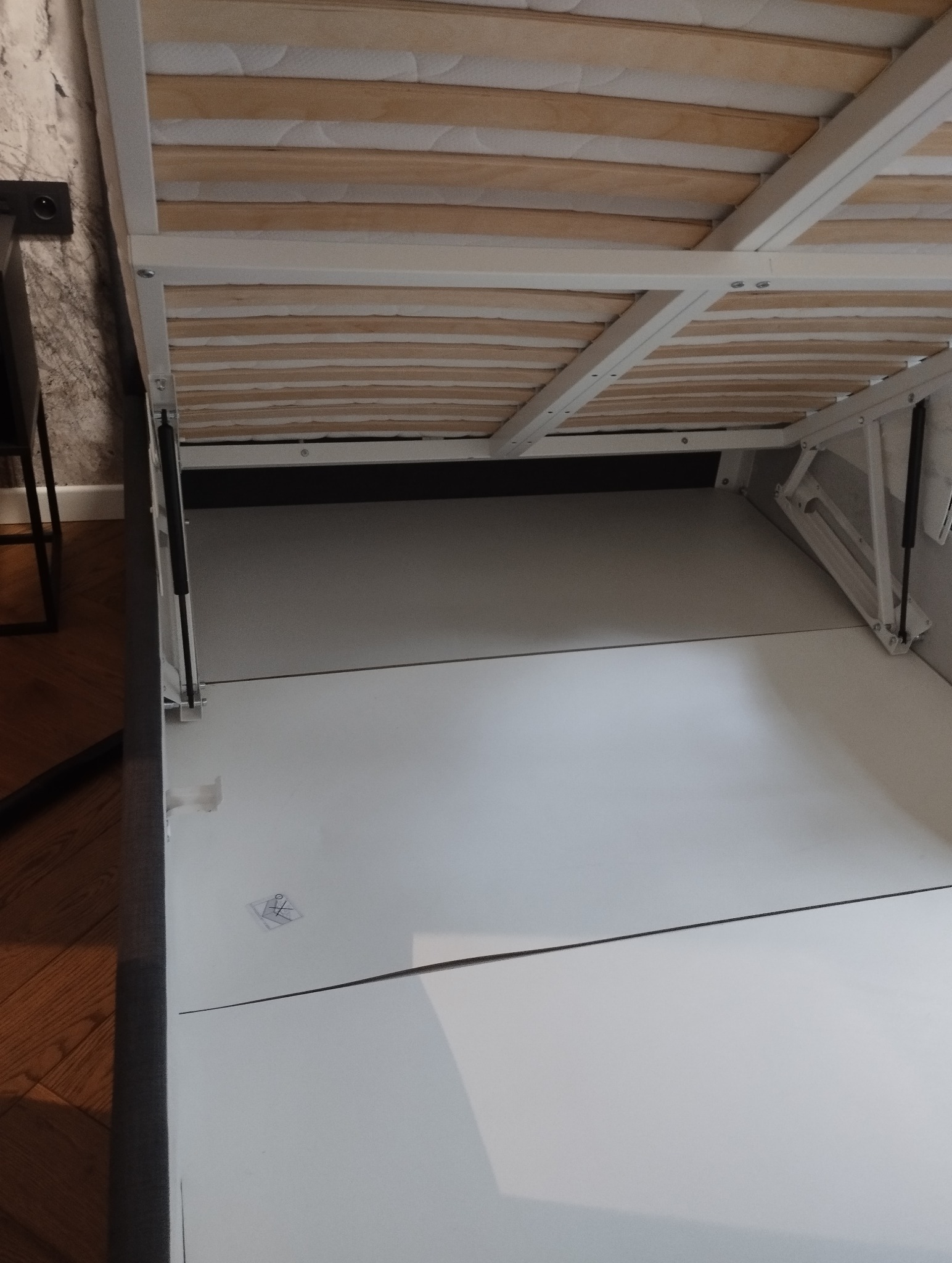 Otwarta rama łóżka z pojemnikiem, widok z dołu. Białe panele pojemnika, drewniany stelaż z materacem. Widoczne mechanizmy podnoszenia.