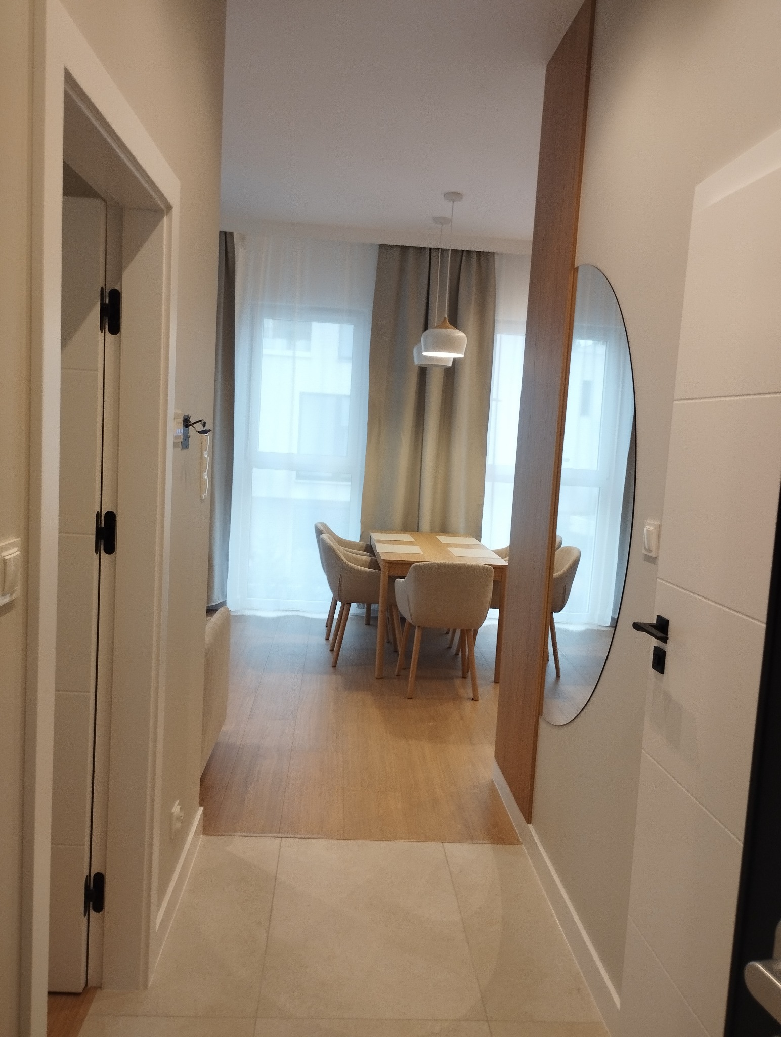 Jasne wnętrze mieszkania z widokiem na stół i krzesła przez otwarte drzwi. Podłoga wyłożona płytkami przechodzi w drewnianą podłogę w dalszej części pomieszczenia.