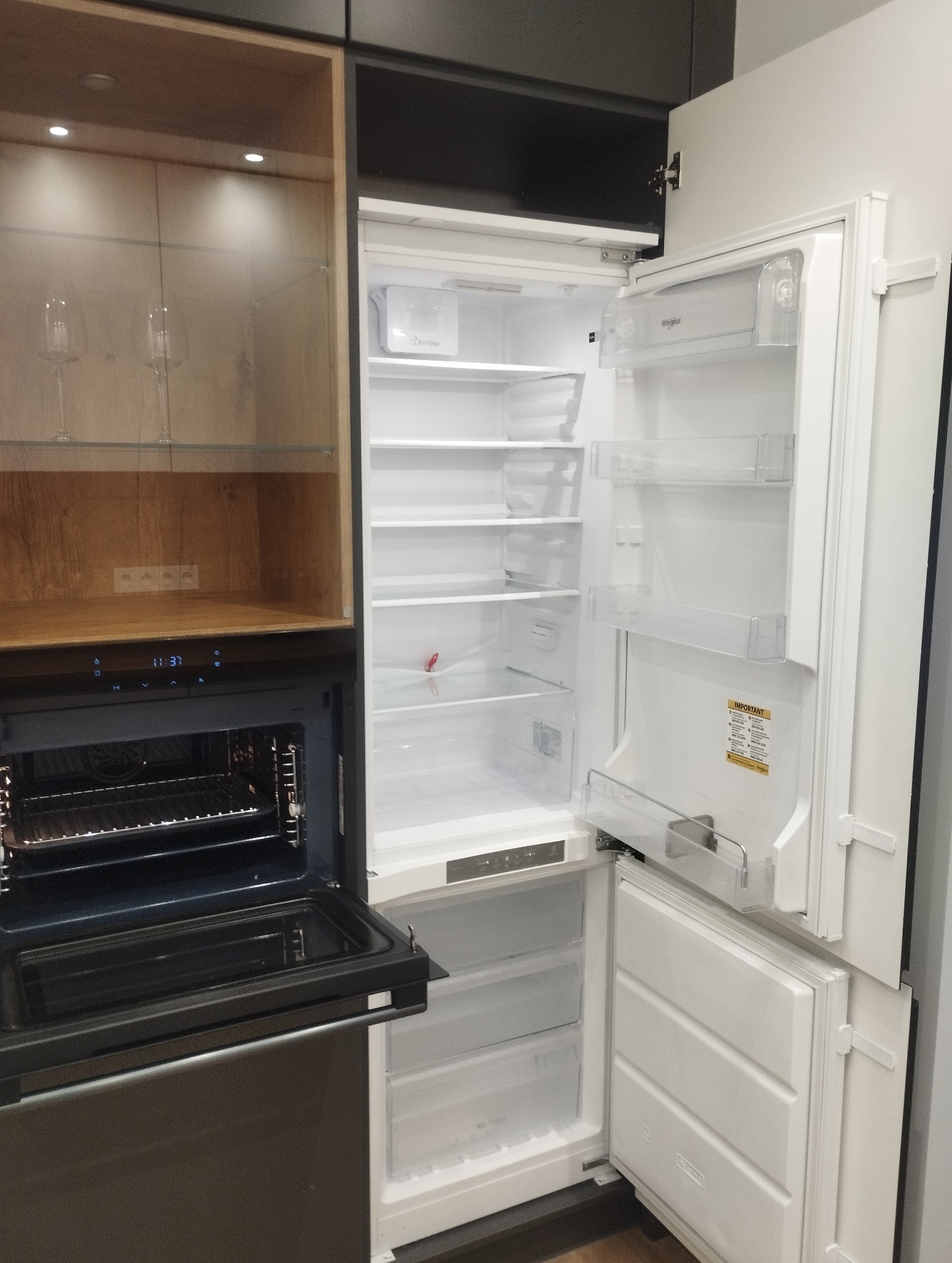 Pusta, otwarta lodówka Whirlpool w zabudowie kuchennej, obok piekarnik i przeszklona szafka. Widoczne półki i szuflady. Nowoczesny design, czyste wnętrze.