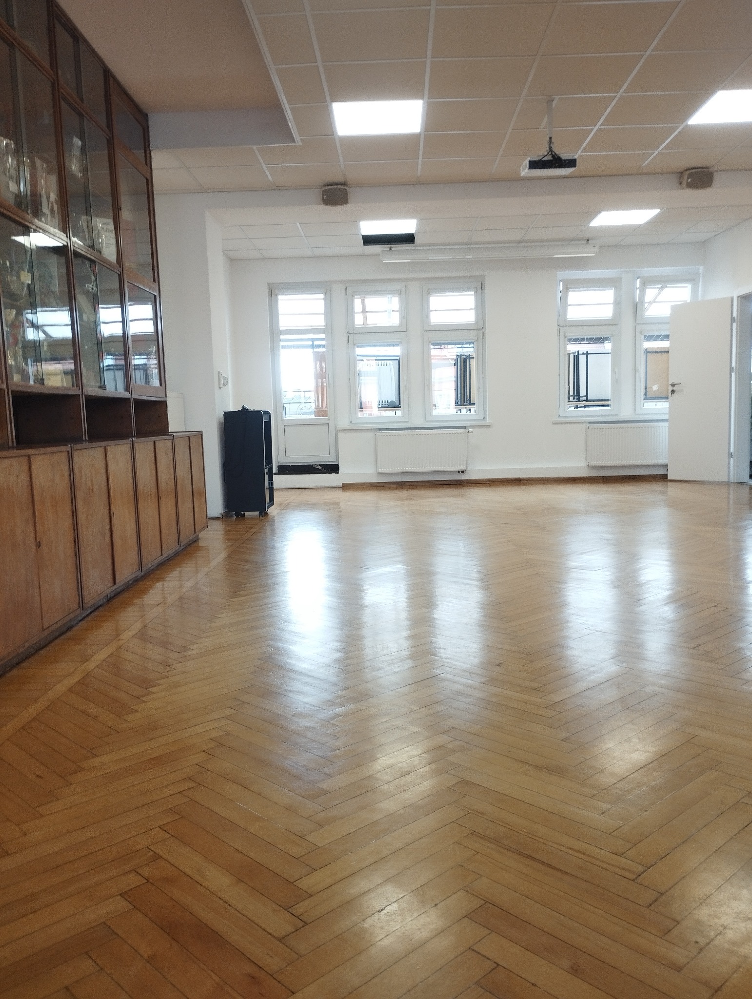 Wnętrze przestronnego pomieszczenia z parkietem ułożonym w jodełkę, po sprzątaniu. Widoczne okna, grzejniki, szafki wzdłuż ściany i projektor pod sufitem.