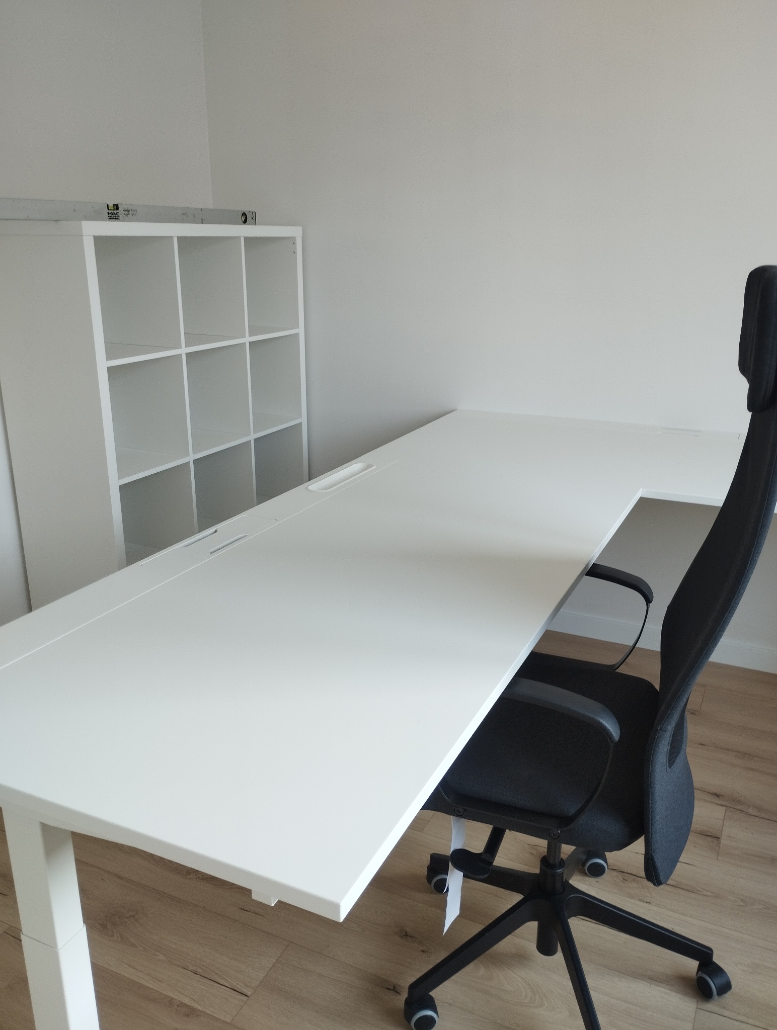 Białe biurko z regulowaną wysokością i czarne krzesło biurowe na tle białej ściany i regału. Minimalistyczne wnętrze biurowe, jasne oświetlenie, drewniana podłoga.