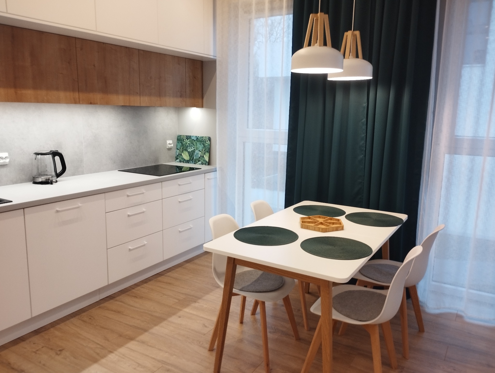 Jasna kuchnia z białymi szafkami i drewnianymi akcentami. Stół z czterema krzesłami i dekoracjami. Nowoczesne oświetlenie i zasłony w tle. Widok na czyste, uporządkowane wnętrze.