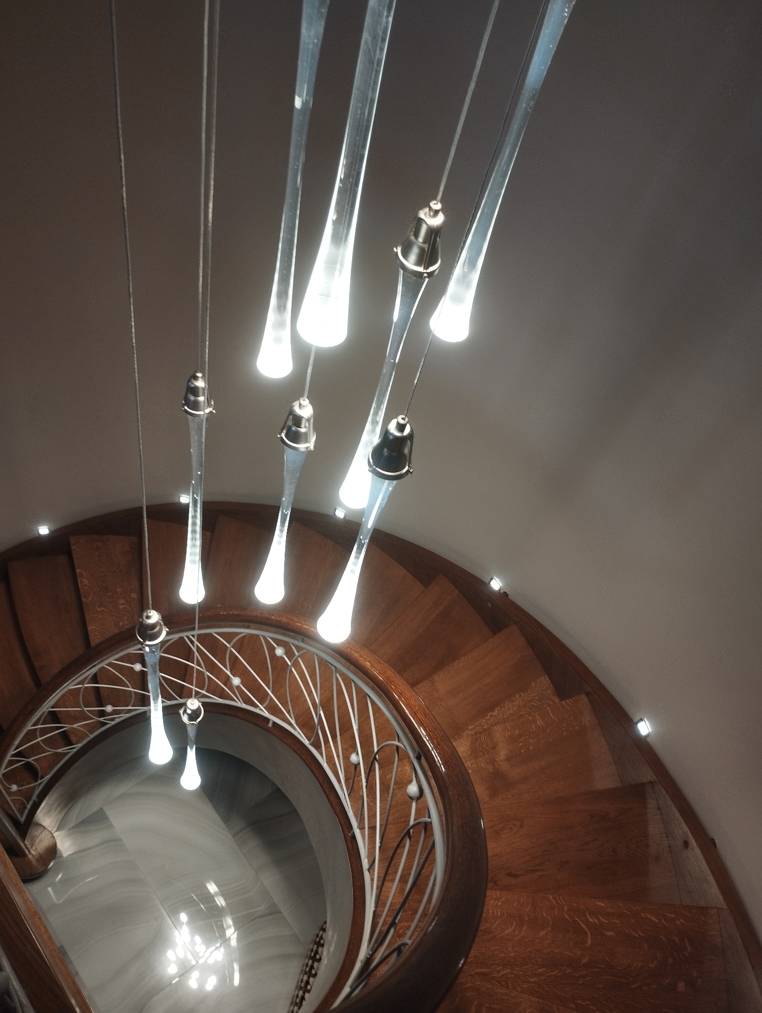 Spiralne schody z drewnianymi stopniami i metalową balustradą, oświetlone wiszącymi lampami z przezroczystego szkła, widziane z dołu, tworzące elegancki efekt wizualny.