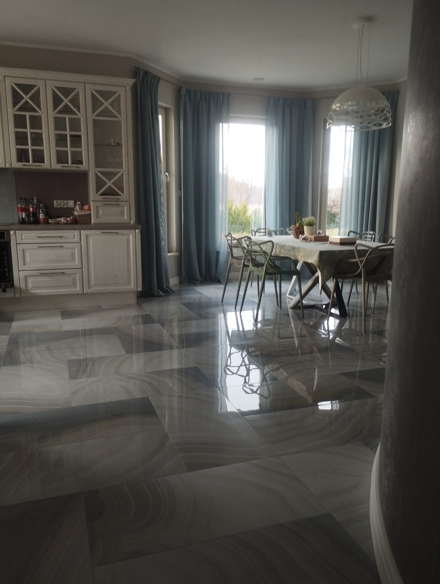 Błyszcząca, szara podłoga z płytek w przestronnej jadalni we Wrocławiu. Widoczne krzesła, stół nakryty do posiłku i okna z niebieskimi zasłonami.