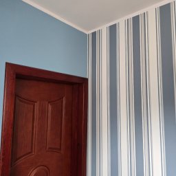 Mateusz Wójtowicz - Fragment wnętrza: brązowe drzwi z ozdobnym frezem, błękitna ściana i ściana w pionowe, szaro-białe pasy. Widok na róg pokoju, z delikatnym oświetleniem.