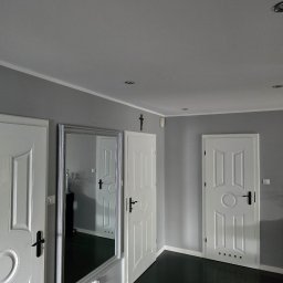 Mateusz Wójtowicz - Szare wnętrze po remoncie: trzy białe, klasyczne drzwi, lustro w srebrnej ramie, ciemna podłoga. Minimalistyczny design z krzyżem nad drzwiami.