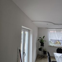 Mateusz Wójtowicz - Jasne, świeżo pomalowane wnętrze z białymi ścianami, widoczne drzwi balkonowe, dekoracyjny sufit i rośliny doniczkowe. Minimalistyczny wystrój.