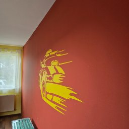 Mateusz Wójtowicz - Czerwona ściana z żółtą naklejką auta w pokoju z oknem i pudłem. Nietypowy detal dekoracyjny w odcieniach czerwieni i żółci.