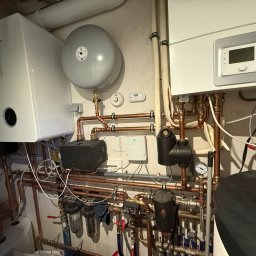 RONA GROUP SPÓŁKA Z OGRANICZONĄ ODPOWIEDZIALNOŚCIĄ - Złożona instalacja grzewcza z kotłem Bosch, zbiornikiem wyrównawczym i licznymi rurami miedzianymi, zaworami oraz filtrami. Widoczne elementy automatyki i sterowania.