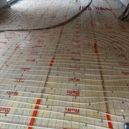 RONA GROUP SPÓŁKA Z OGRANICZONĄ ODPOWIEDZIALNOŚCIĄ - Białe rury ogrzewania podłogowego ułożone spiralnie na izolacji z czerwonym nadrukiem Roth, widok z góry, fragment podłogi z rurami poza kadrem.