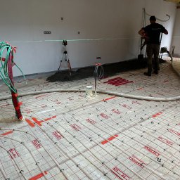 RONA GROUP SPÓŁKA Z OGRANICZONĄ ODPOWIEDZIALNOŚCIĄ - Układanie ogrzewania podłogowego Roth z użyciem poziomicy laserowej. Widoczny pracownik w trakcie wylewania posadzki. Rury grzewcze zamocowane do podłoża.