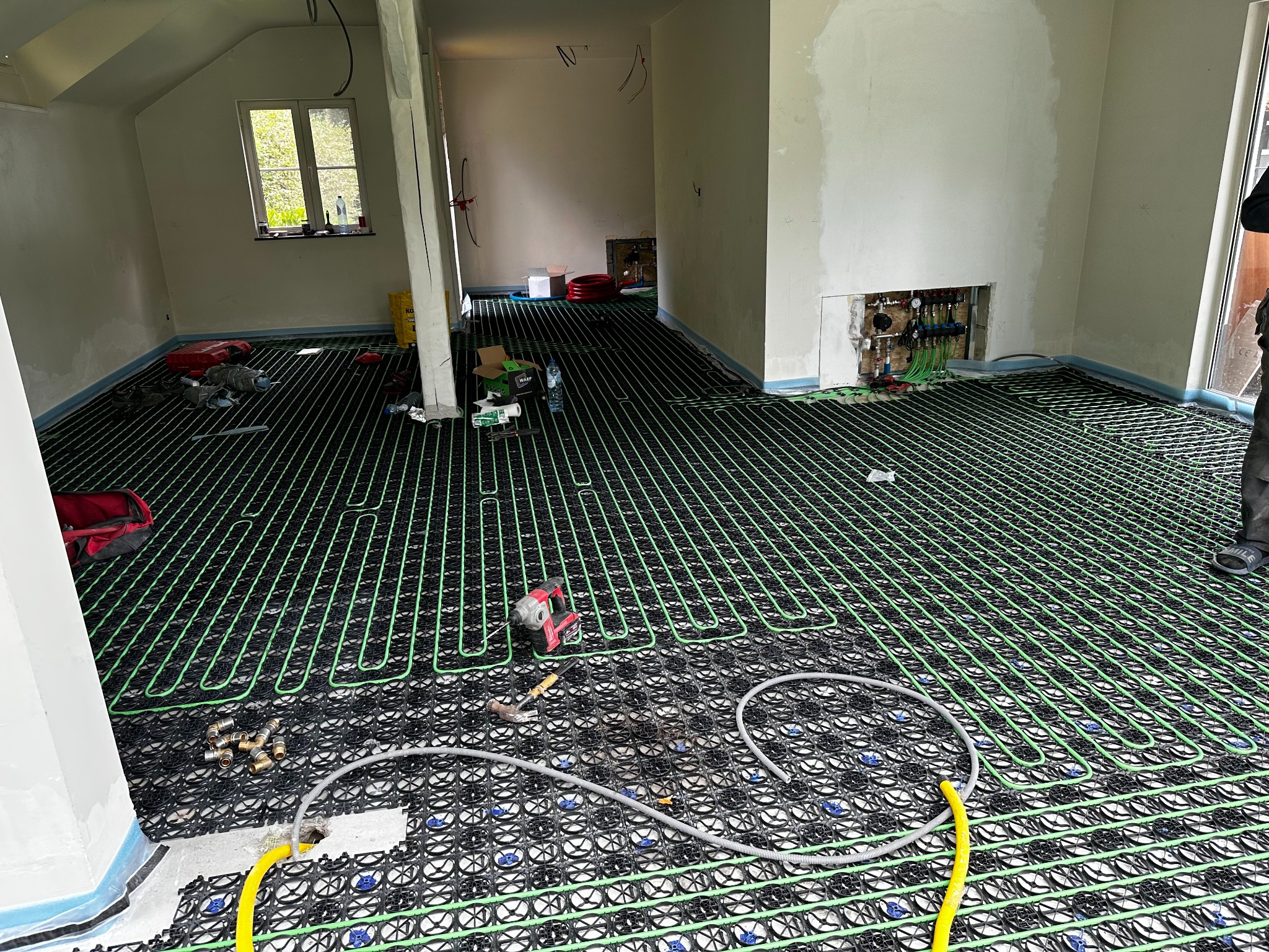 Instalacja ogrzewania podłogowego w trakcie montażu: zielone rurki ułożone na czarnej macie z narzędziami. Widoczny rozdzielacz i fragment instalacji w ścianie.
