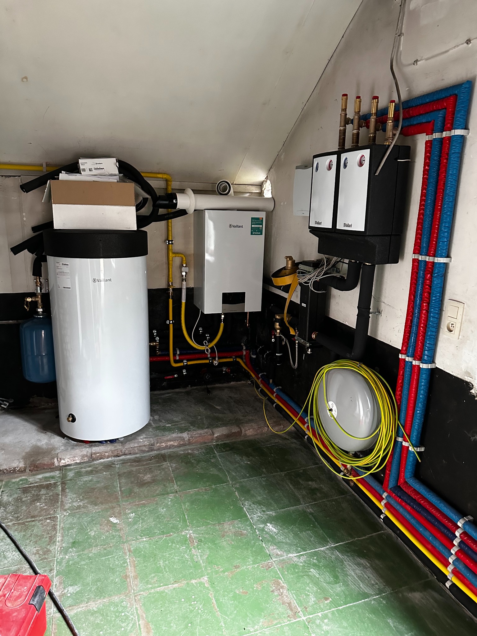 Zmontowana instalacja grzewcza Vaillant w kotłowni: biały zasobnik, piec, czarne moduły, rury w izolacji czerwonej i niebieskiej, żółte rury gazowe. Zielona, popękana podłoga.