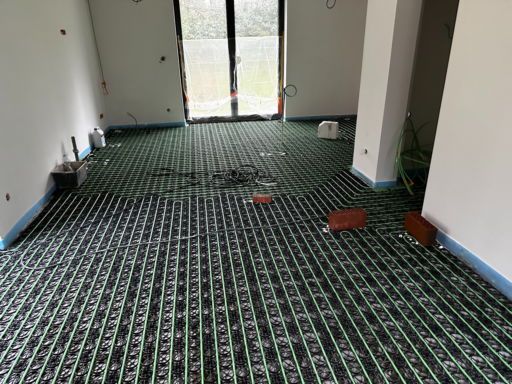 Ułożona instalacja ogrzewania podłogowego w jasnym pomieszczeniu z dużym oknem. Widoczne rury w kolorze zielonym i czarnym, cegły obciążające instalację.