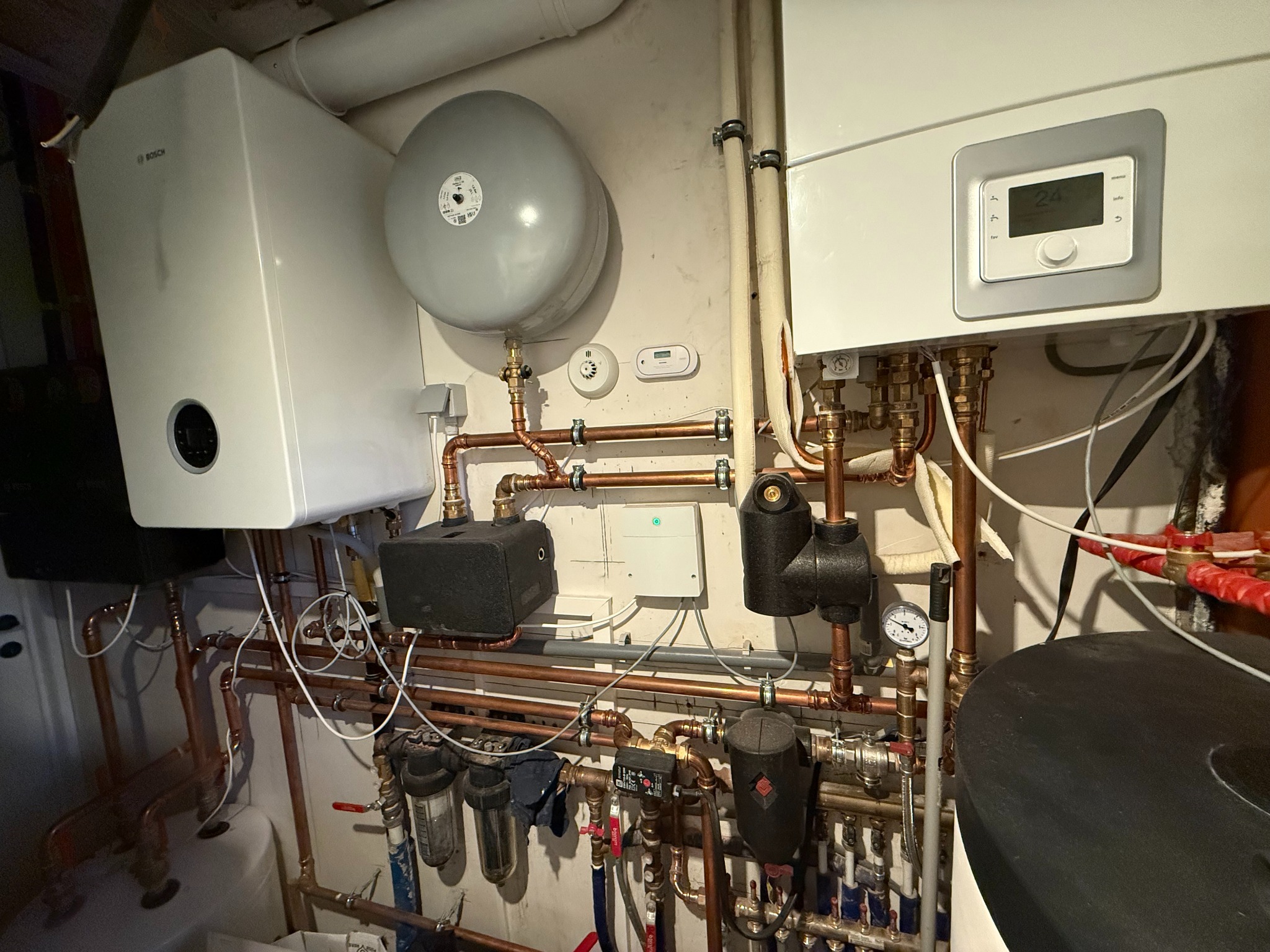 Złożona instalacja grzewcza z kotłem Bosch, zbiornikiem wyrównawczym i licznymi rurami miedzianymi, zaworami oraz filtrami. Widoczne elementy automatyki i sterowania.