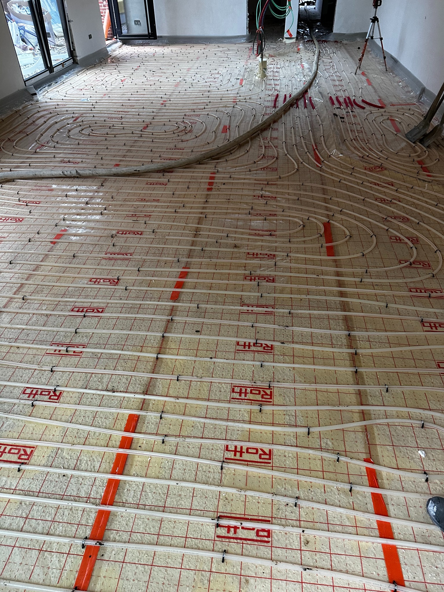 Białe rury ogrzewania podłogowego ułożone spiralnie na izolacji z czerwonym nadrukiem Roth, widok z góry, fragment podłogi z rurami poza kadrem.